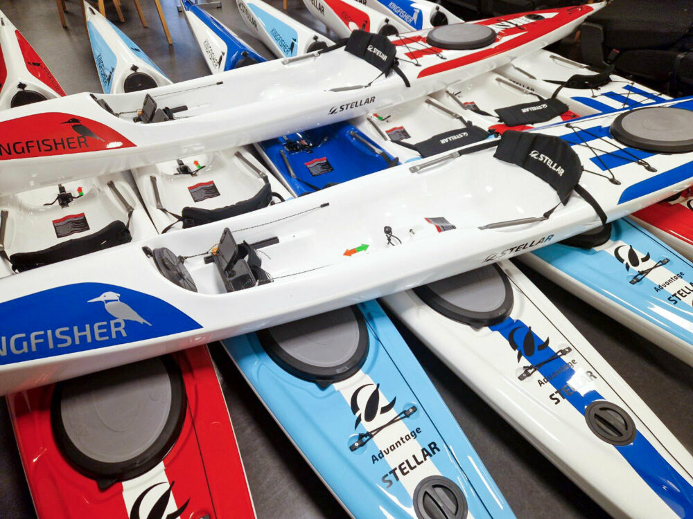 Foto: Kajakker i flere farger ligger i to lag, underst ligger de fem stykker og på tvers over ligger det to. Fargene er rødt, hvitt, blått og lyseblått.