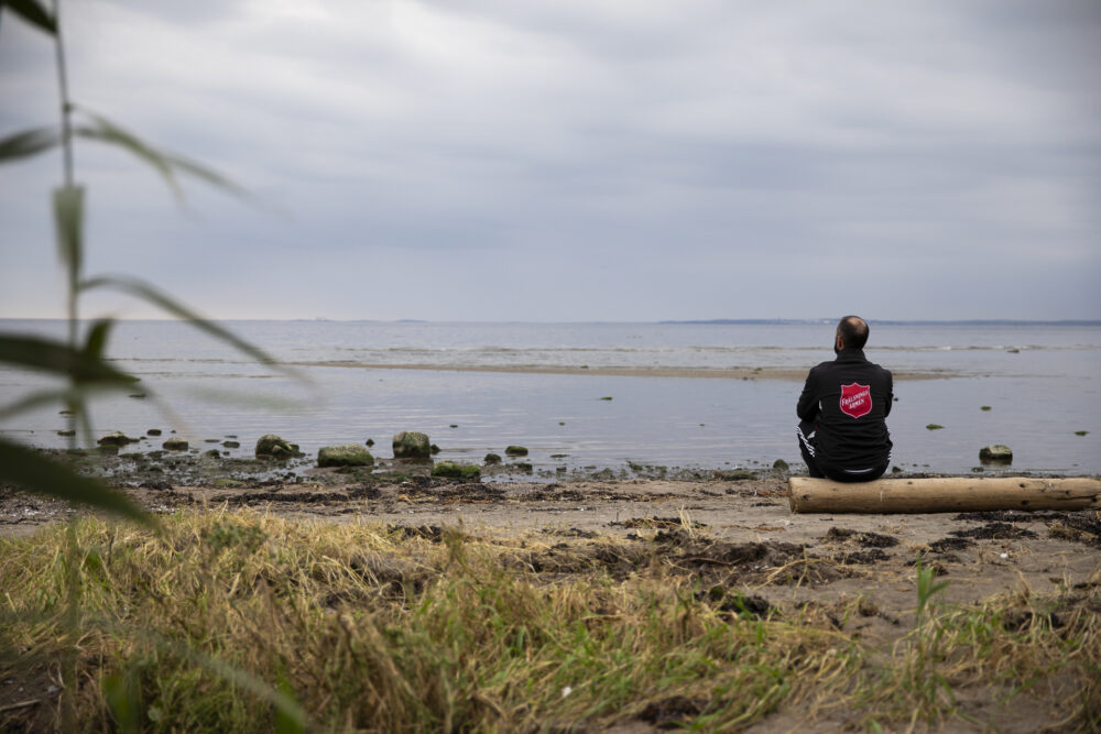 Foto: Mann med jakke med stor logo fra Frelsesarmeen på ryggen. Han sitter på en tømmerstokk og ser utover havet på en øde strand. Været er overskyet.