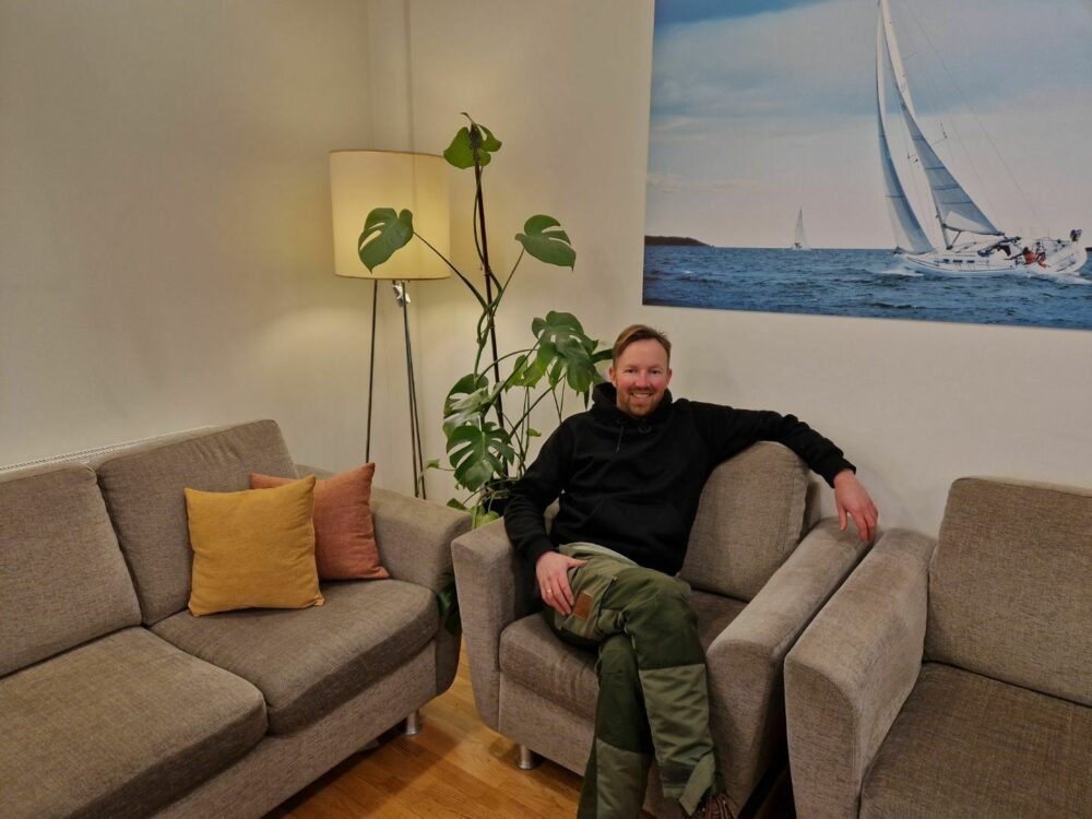 Foto: Smilende mann tidlig i 40-årene sitter i turbukse og genser i en diger, grå lenestol. Han har mørkeblondt hår og skjegg.