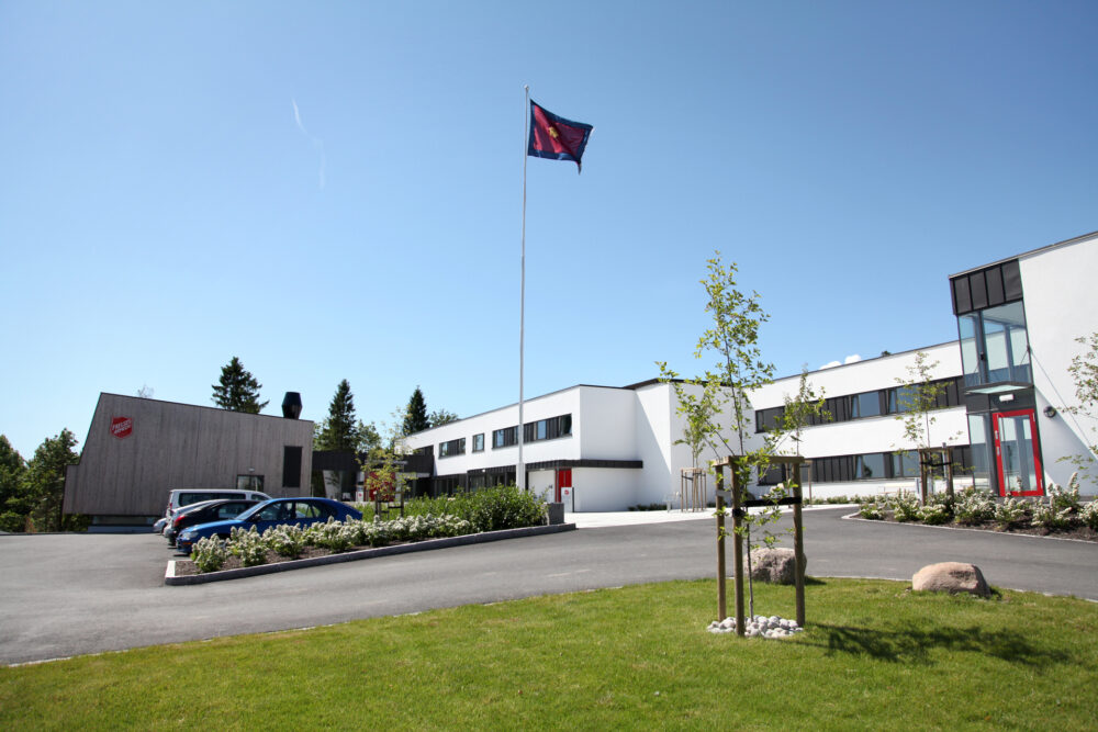 Foto: Hvitt, moderne funkisbygg med flagg fra Frelsesarmeen i toppen av flaggstanga og bilder på parkeringsplassen utenfor.
