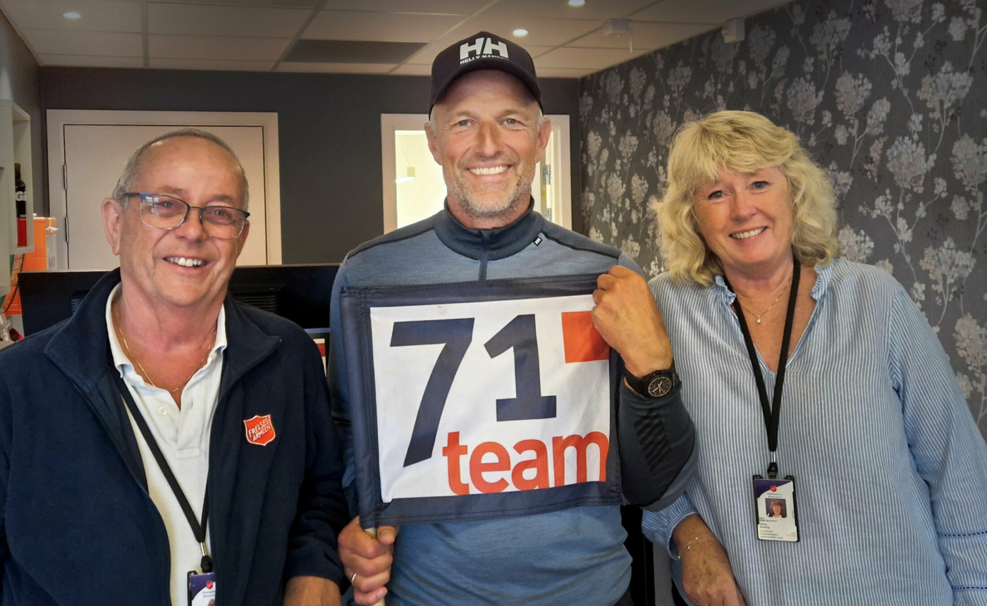 Foto: To menn og en kvinne i 50- og 60-årene smiler til kamera fra en resepsjon med blomstrete tapet på veggen bak. Mannen i midten er mye høyere enn de andre to og har på mørk caps og et stykke tøy med logoen til 71 grader nord team på.