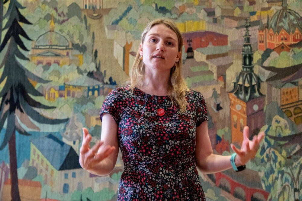 Foto: Kvinne i 30-årene i blomstrente kjole snakker. Gestikulerer med hendene. I bakgrunnen er det et fargerikt, vevd teppe med motiv fra kvartalene rundt Oslo domkirke.