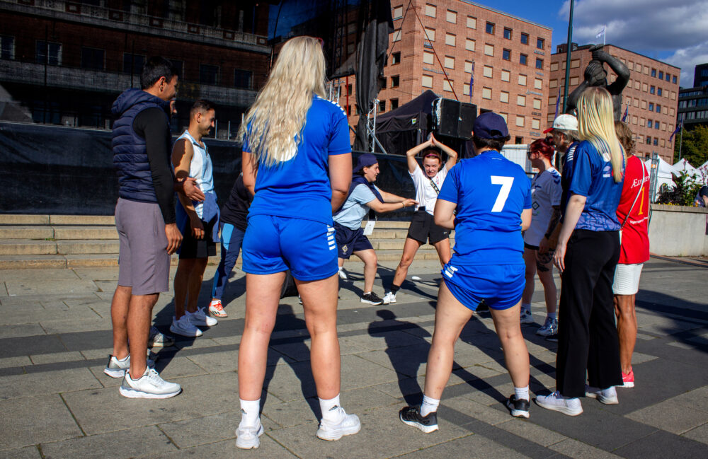 Foto: En gruppe mennesker står i ring og holder på med en lek. En av dem, en kvinne i hvit T-skjorte, holder armene over hodet. I bakgrunnen er det rød mursteinsbygg på 8-10 etasjer.