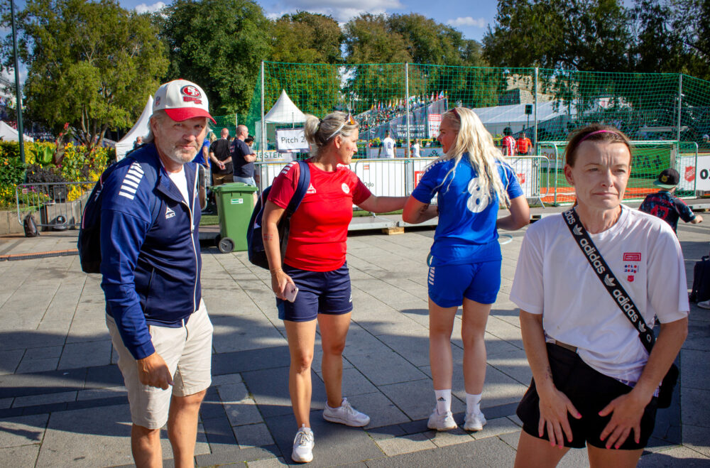 Foto: Fire personer i treningstøy, en av dem er mann. De står ute på en plass der det er fotballbinger rundt og grønne trær.