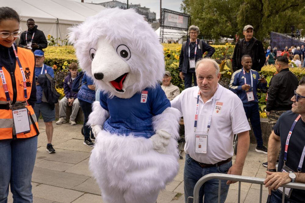 Foto: Mann med grått hår i 60-årene støtter en maskot i ryggen og hjelper rpå vei. Rundt er det sperringer og mye folk, bl.a. en vakt i oransje vest.