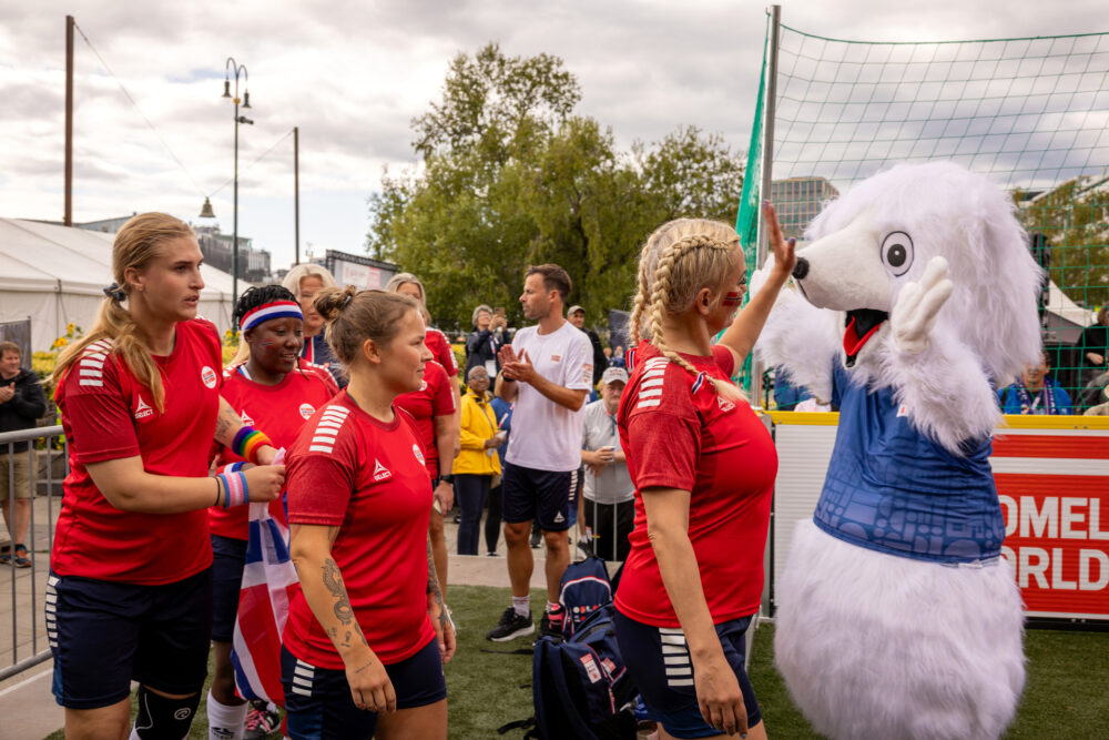 Foto: En hvit isbjørn-maskot med blå trøye gir high five til et kvinnelag i røde T-skjorter med norske flagg. De kommer på rekke inn på banen.