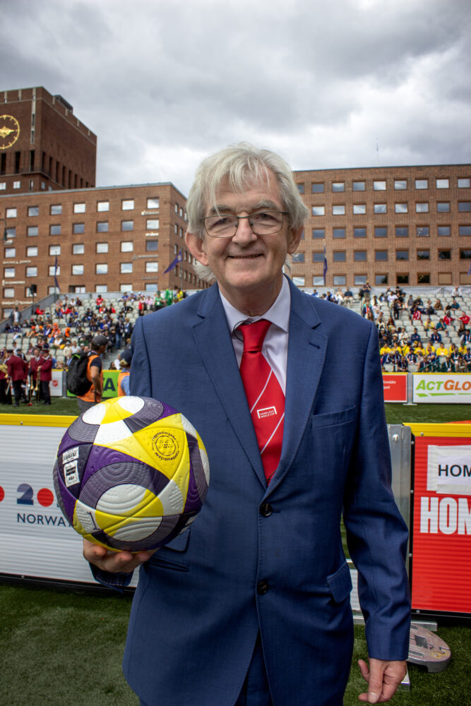 Foto: Mann i 70-årene med grått hår og briller står ie ne blå dress. Han har på seg et rødt slips og holder en gul, hvit og blå fotball i høyre hånd. Han står på en liten fotballbane på Rådhusplassen med mange folk på tribunene.