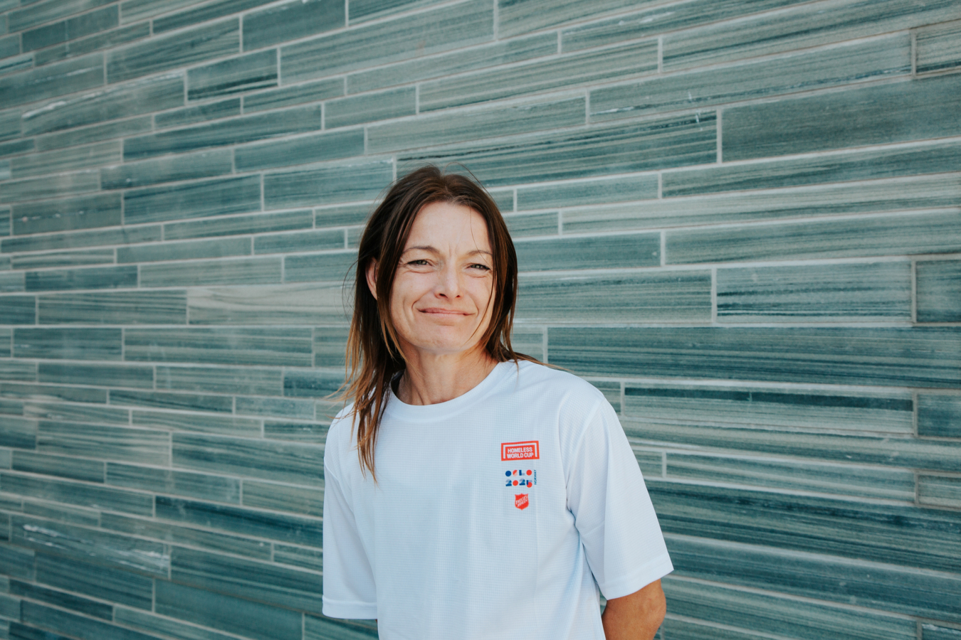 Foto: Kvinne i hvit T-skjorte der det står Homeless World Cup Oslo 2025. Hun har halvlangt, mørkt hår og smiler står foran en grå murvegg.