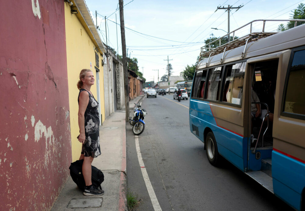 Foto: Norsk kvinne i 50-årene står i en svart og hvit sommerkjole uten ermer i en gate i Latin-Amerika. En lokal liten buss står parkert med døra åpen.