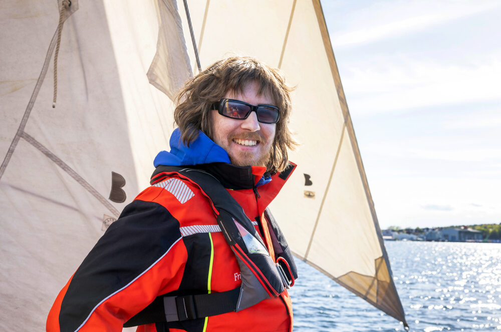 Foto: Ung mann med mørkt hår og skjegg smiler foran seilet. han har på seg rød jakke og solbriller.