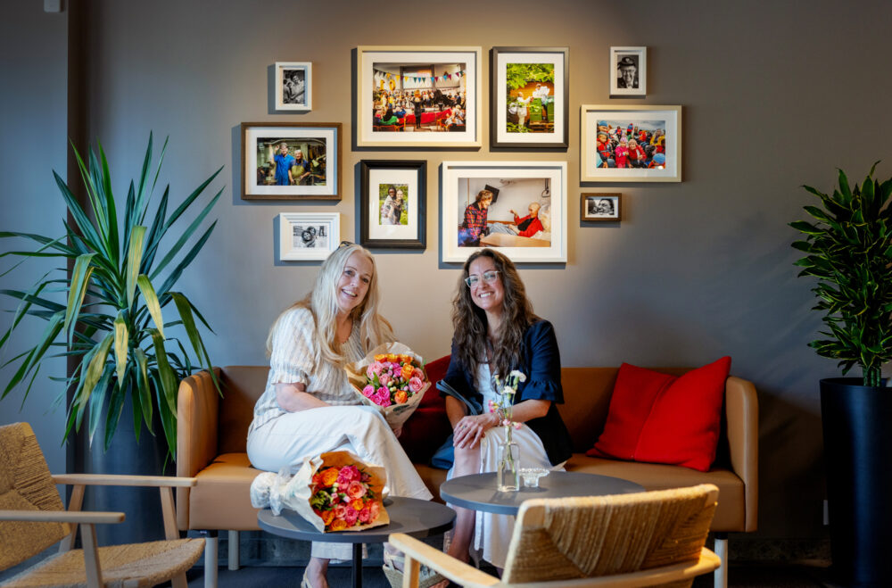 Foto: En blond og en mørkhåret kvinne med briller sitter i en sofa. De har fått hver sin blomsterbukett som takk for innsatsen.