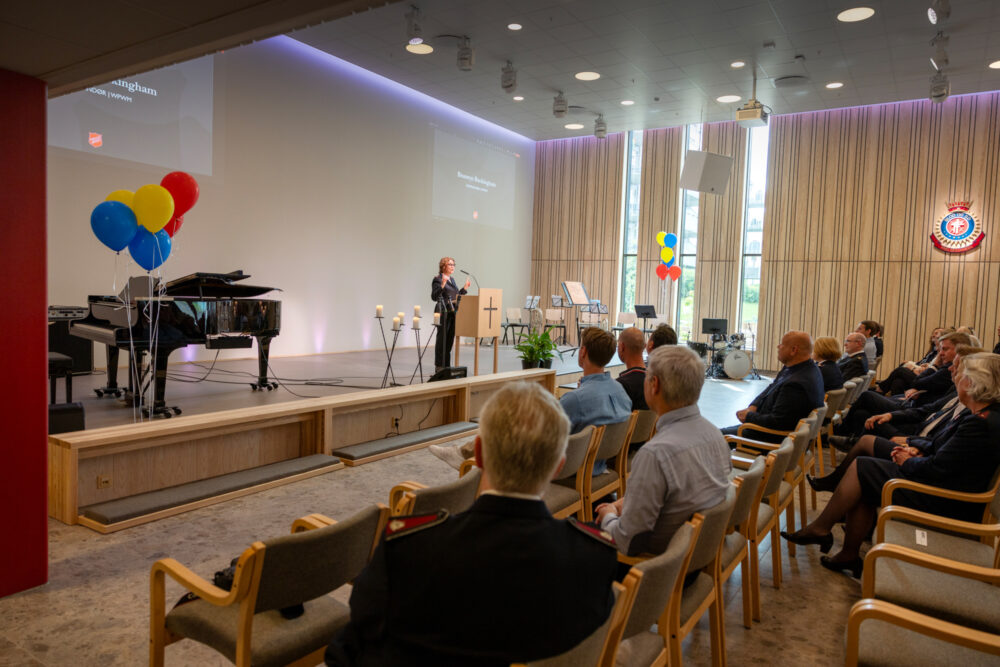 Foto: Kvinne med krøller og briller står på talerstolen. Rundt om er det ballonger. I salen sitter folk på rekke og rad.
