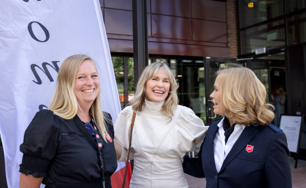 Foto: Tre blonde kvinner i 50-årene står tett sammen utenfor et kontorbygg med en "flagg" der det står velkommen.