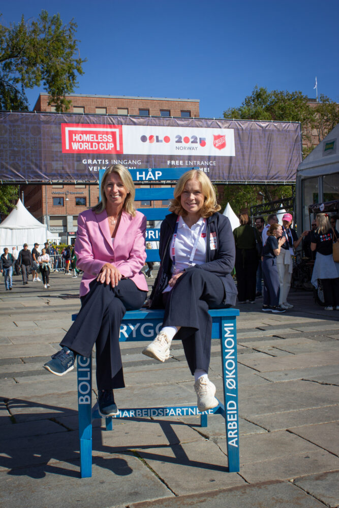 Foto: To blonde kvinner i 40-årene sitter på en kjempestor blå stol ute på Rådhusplassen. I bakgrunner ser vi røde murbygg og et stort banner der det står logoen til Homeless World Cup etterfulgt av ny logo der det stpår Oslos 2025 Norway og så den røde skjoldet til Frelsesarmeen.