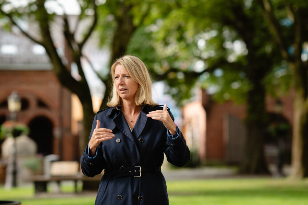 Foto: Kvinne med halvlangt blondt hår i blå frakk gestikulerer utendørs foran noen høye, gamle trær og en rød murbygning.