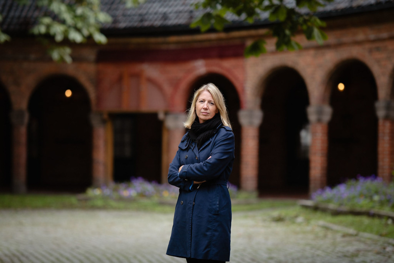 Foto: Kvinne i 40-årene med halvlangt, blondt hår står foran et rødt, stort mursteinsbygg med en rekke bueganger. Hun har på seg blå kåpe og svart skjerf.
