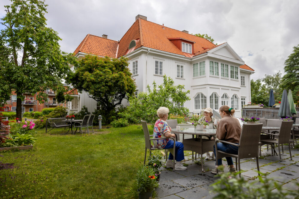 Foto: Tre damer sitter ved et hagebord som står på noen heller i en stor og frodig hage. I bakgrunnen sees en hvit, ærverdig villa med rødt tak.