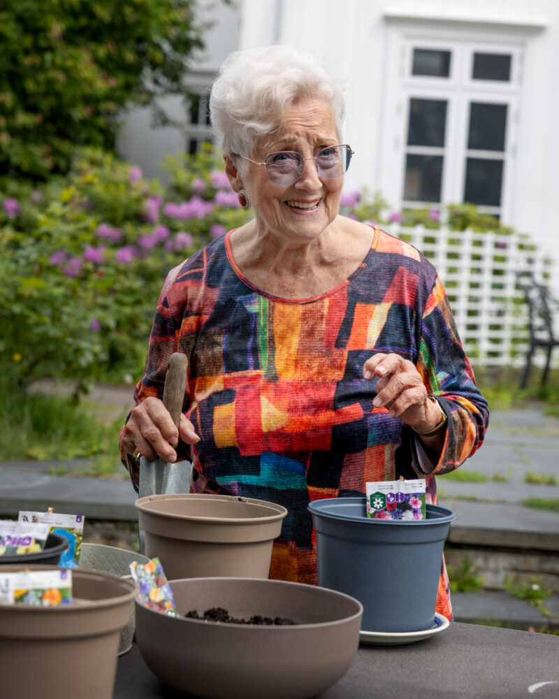 Foto: Eldre kvinne i 80-årene som står ved noen potter der hun har sådd planter.