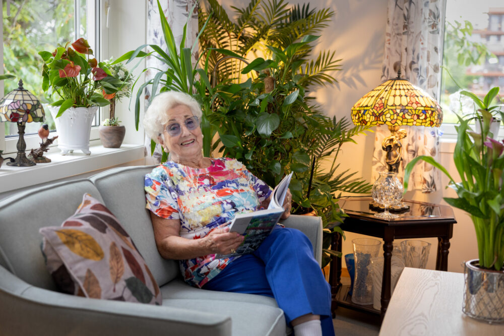 Foto: Eldre kvinne i fargerik bluse sitter i en sofa. Bak henne er det mange planter og en Tiffany-lampe.
