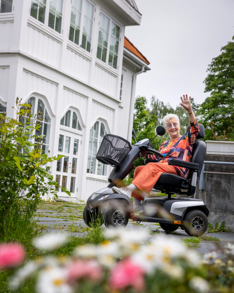 Foto: Eldre kvinne på firhjuling utenfor et hvitt, gammelt hus.