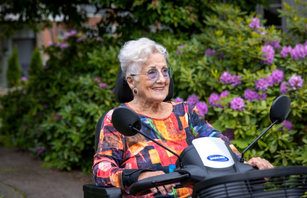 Foto: Kvinne med grått hår og briller med lilla glass sitter på en firhjuling og holder i styret. Hun har på seg fargerik topp.