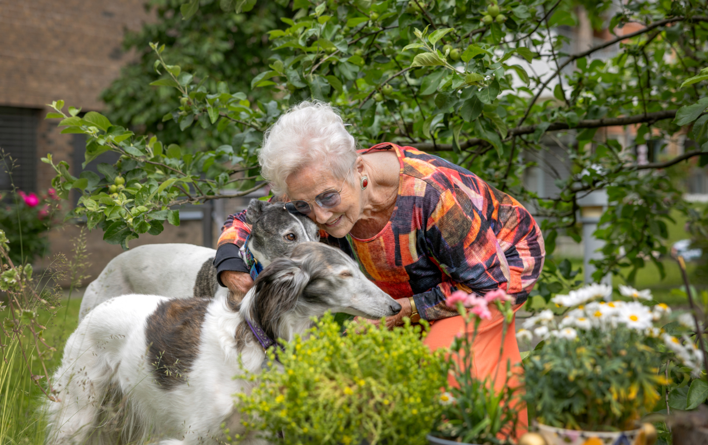 Foto: Eldre kvinne i mønstrrete topp og oransje bukser er i hagen og kose med to under.