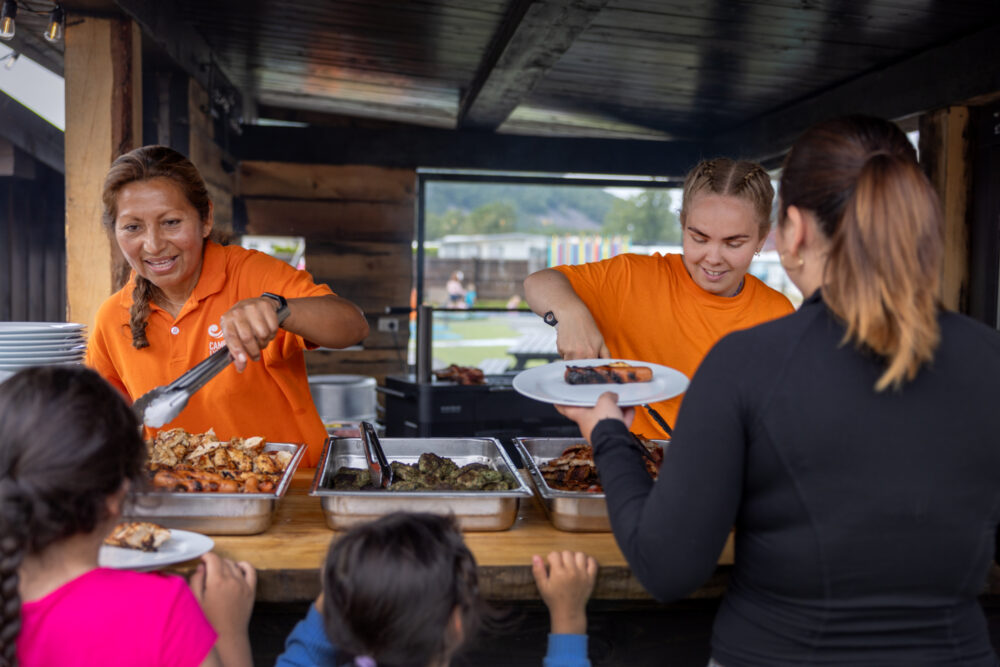 To foto: Det første viser en dame fra Sør-Amerika og en blond jente med fletter, si en grillhytte, om serverer kylling og annet kjøtt til en dame og to jenter med ryggen til kamera. Det andre bildet viser en voksen hånd som gir en tallerken med poteter og to pølser, og to barnehender som mottar.