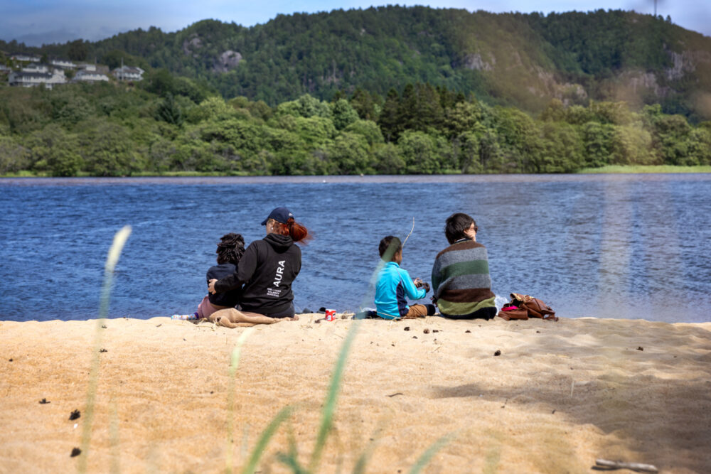 Foto: To voksne og to barn sitter påkledd på en strand med utsikt over på land på den ander siden. Det er grønt og frodig.