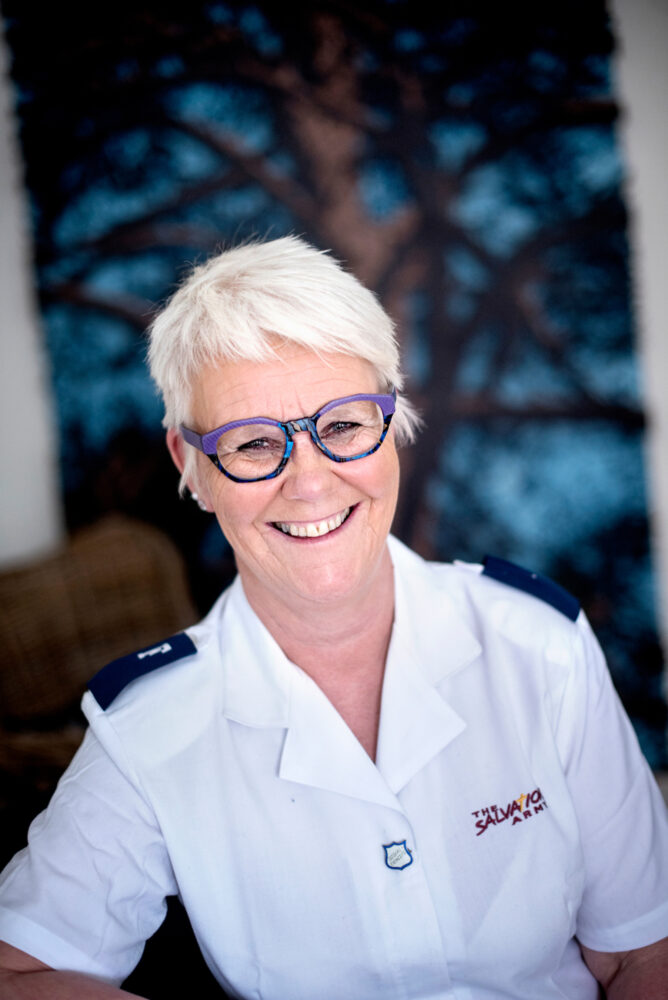 Foto: Moden kvinne i hvit uniformsskjorte smiler med fargerike briller. Bak på veggen er et stort maleri i mørke farger.