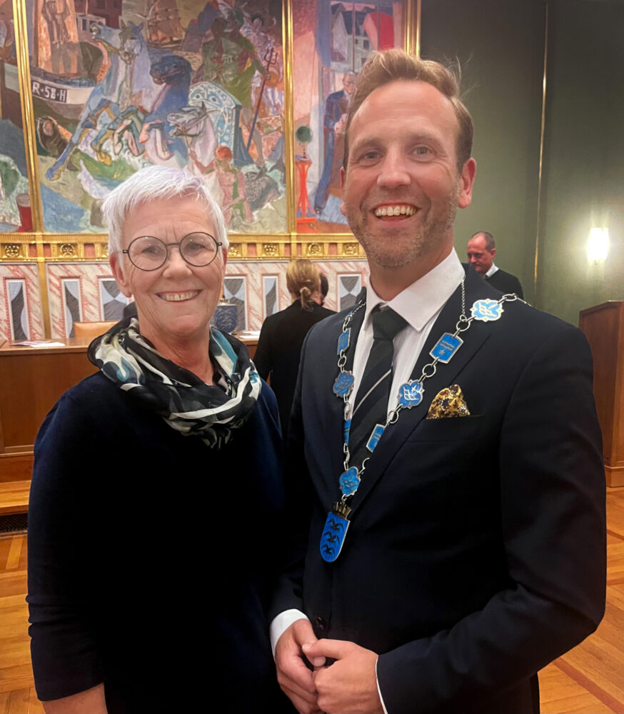 Foto: Ung mann i dress med ordførerkjede rundt halsen står ved siden av en voksen dame med briller og pene klær. De står i en stor, utsmykket sal.