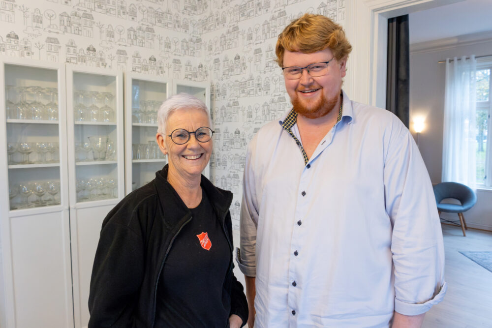 Foto: Moden kvinne i svart jakke med logo fra Frelsesarmeen sammen med yngre, høy mann med briller og skjegg inne i en finstue.