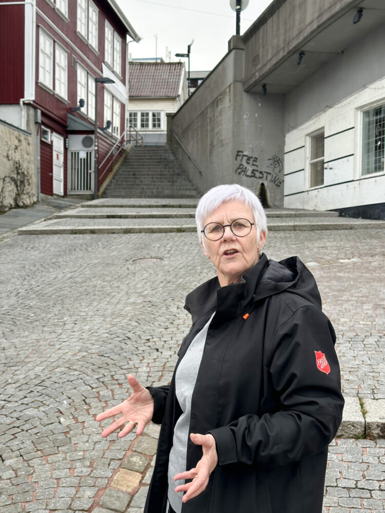 Foto: Samme dame i mørk jakke fra Frelsesarmeen står i enden av en brosteinsbelagt bakke. Til høyre ser vi starten av ei bru, til venstre og rett fram gamle hus i et gate.