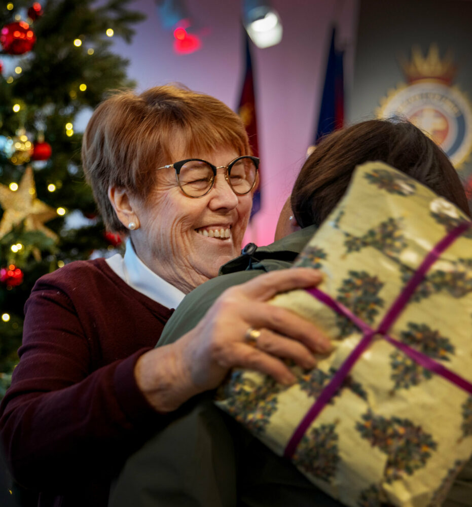 Foto: Eldre kvinne fra Frelsesarmeen gir en kvinne en klem. I hånden holder hun en gave som hun skal gi til henne. I bakgrunnen står et pyntet juletre og flagg fra Frelsesarmeen sammen med kronen på veggen (symbol i Frelsesarmeen).