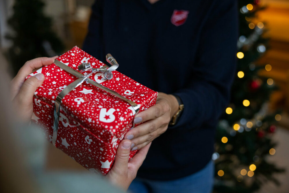 Foto: Kvinnehender gir en innpakket julegave til et par andre kvinnehender. Hun som gir har på seg en genser med logo fra Frelsesarmeen.