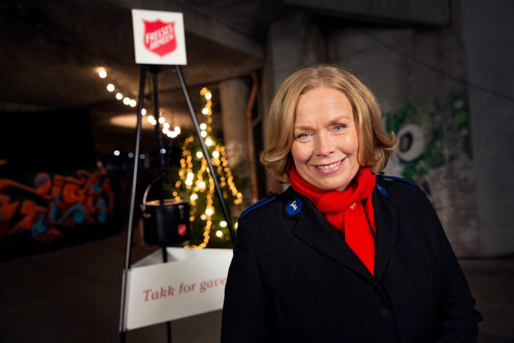 Foto: Kvinne med rødt skjerf og mørk uniformsjakke fra Frelsesarmeen står foran et stativ med en julegryte fra Frelsesarmeen.