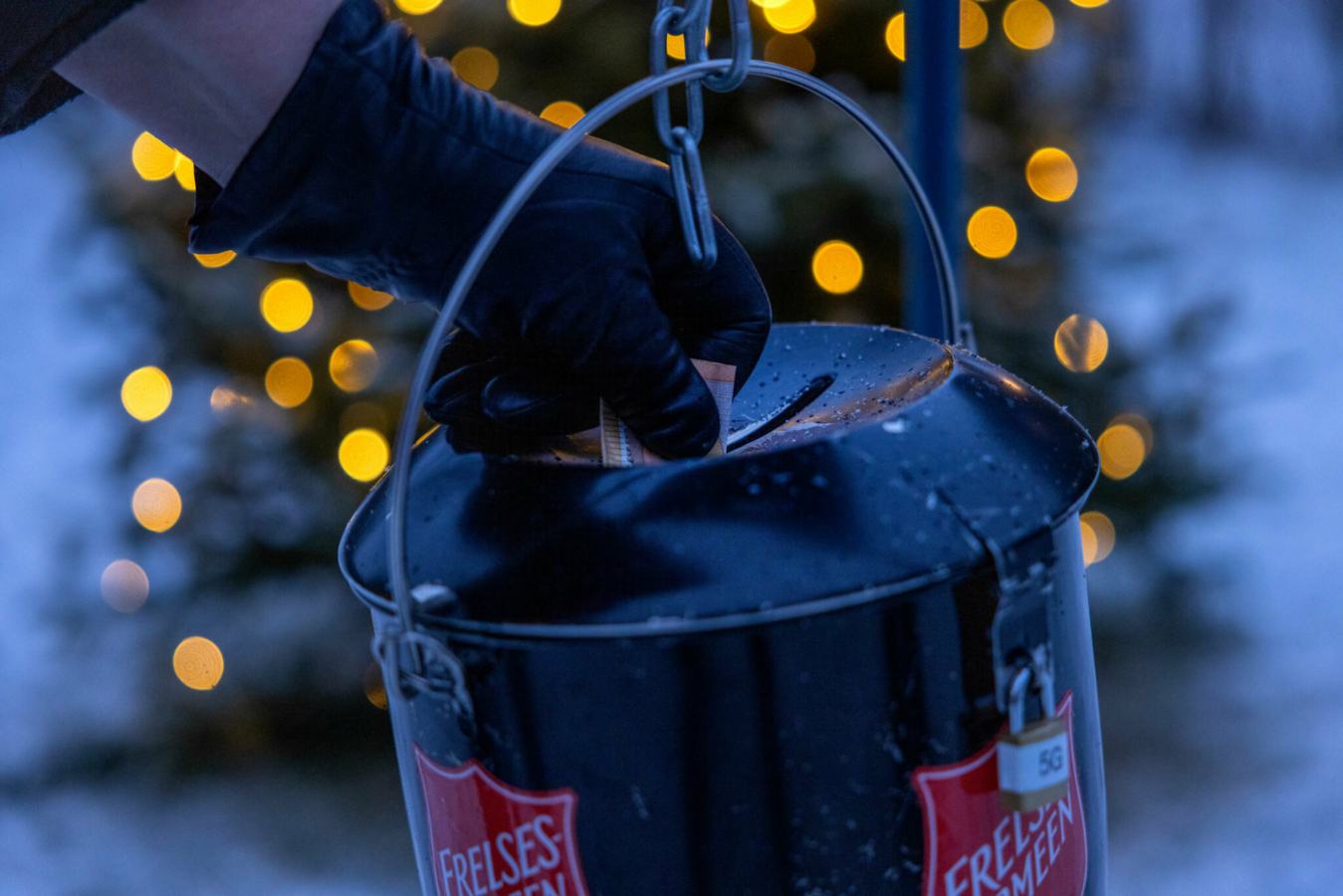 Foto: Hånd med svart skinnhanske som putter en seddel i en julegryte. Det skjer utendørs med snø og julelys på et tre like bak.