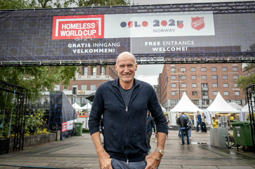 Foto: Mann i 60-årene med blank isse står ute på en åpen plass. Bak ser vi rådhuset i Oslo og et banner der det står Homeless World Cup Oslo 2025 Norway og logoen til Frelsesarmeen.