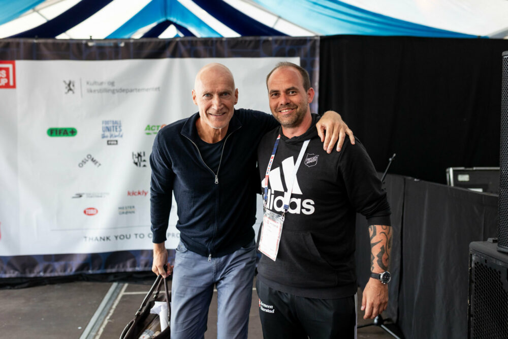 Foto: To menn i 60- og 40-årene holder armen rundt hverandre inne i et stort telt. De har på seg idrettstøy.