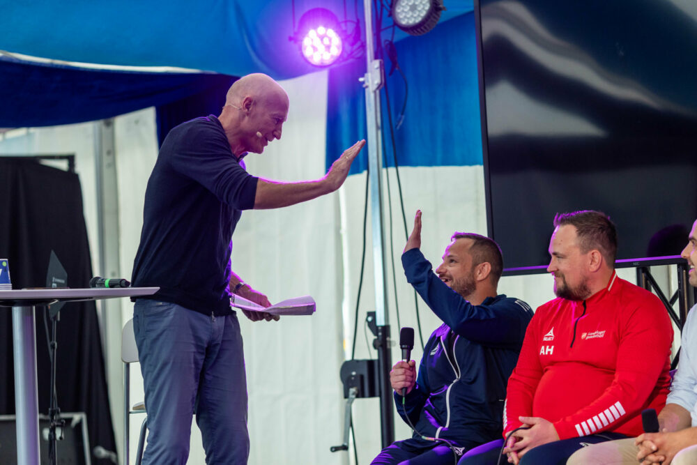 Foto: Mann i 60-årene med blank isse gir en high five til en fotballspiller som sitter på rekke i et panel. De sitter i et stort, utendørs telt.