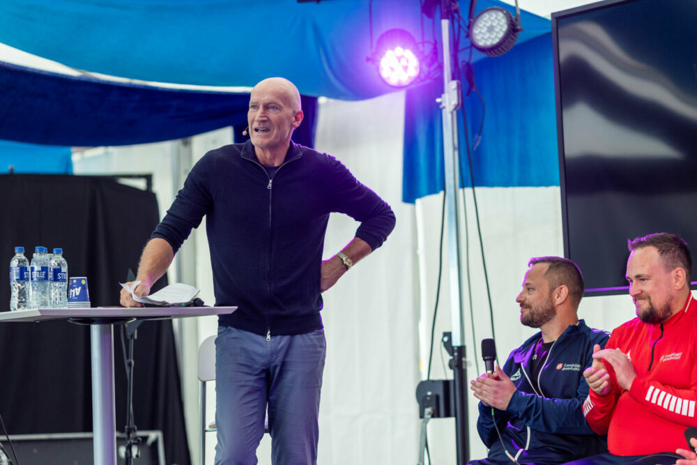 Foto: Mann i 60-årene med blank isse står ved et bord på en scene. Bak ham sitter noen menn i treningstøy på rekke.