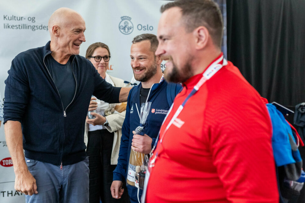 Foto: Mann i 60-årene med blank isse legger skulderen på en gatefotballspiller i blå treningsjakke. Ved siden av ham står en annen mann i rød jakke.