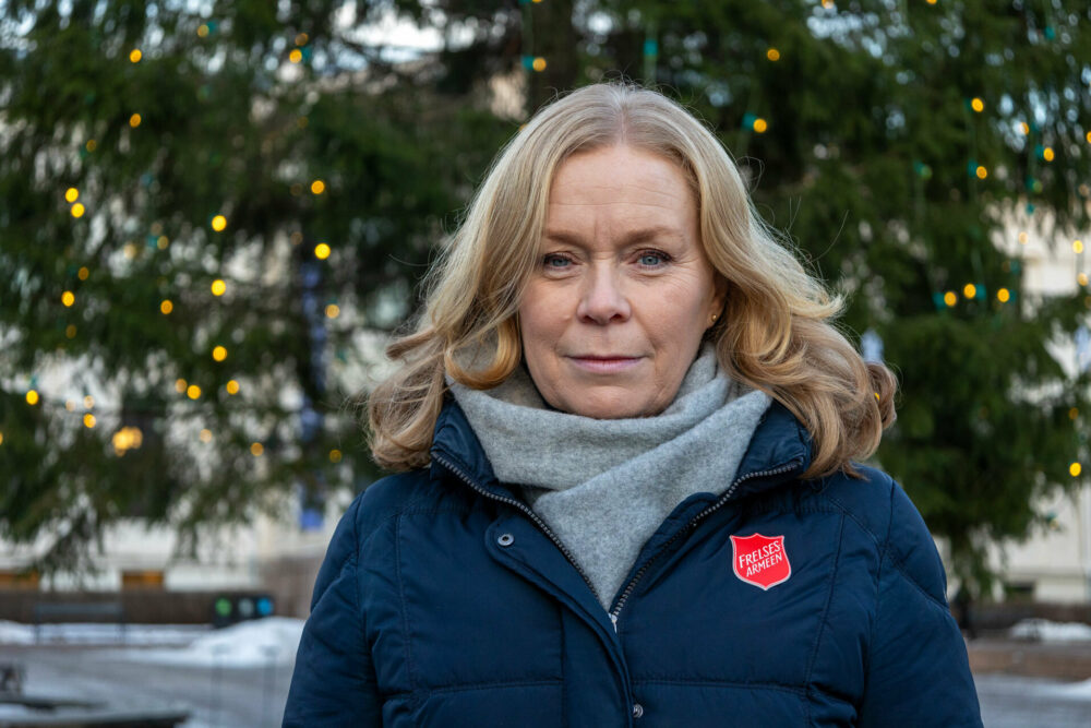 Foto: Kvinne med gyllenblondt hår står foran en stor julegran med lys ute. Hun har på seg blå ytterjakke med logo fra Frelsesarmeen og grått skjerf i halsen.