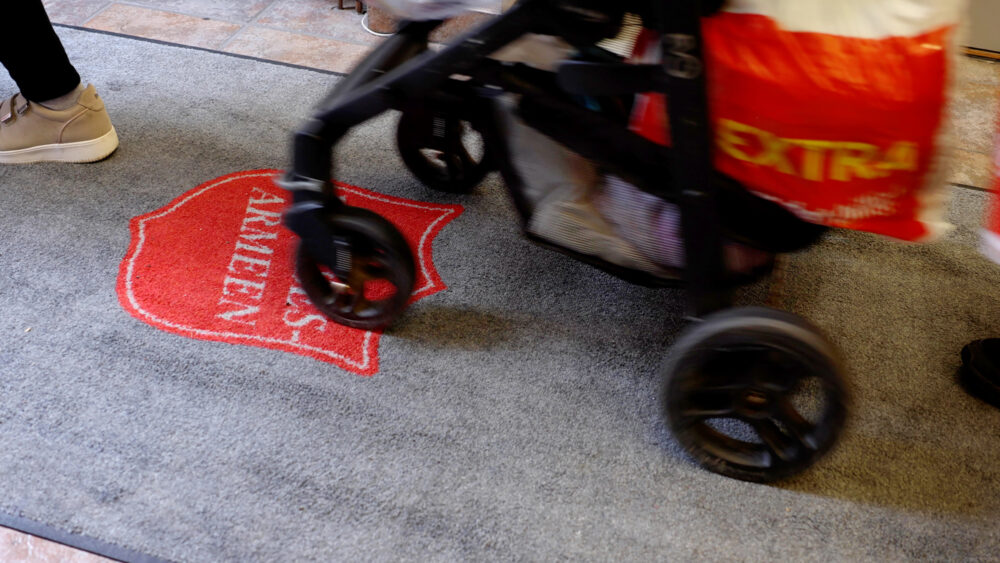 Foto: En barnevogn som ruller ever en grå løper med skjoldet til Frelsesarmeen på. På vogna henger det en pose med matvarer. Vi ser ingen mennesker, kun føtter med sko.