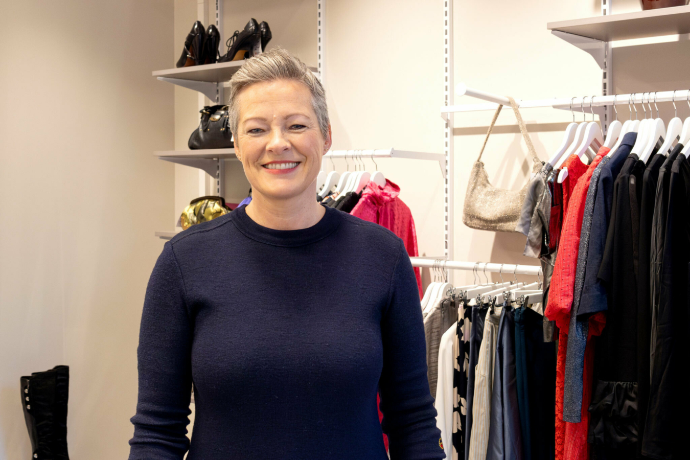 Foto: Kvinne i 50-årene med kort, grått hår og blå genser står i en bruktbutikk. Bak henne er det hyller med klær, sko og vesker.