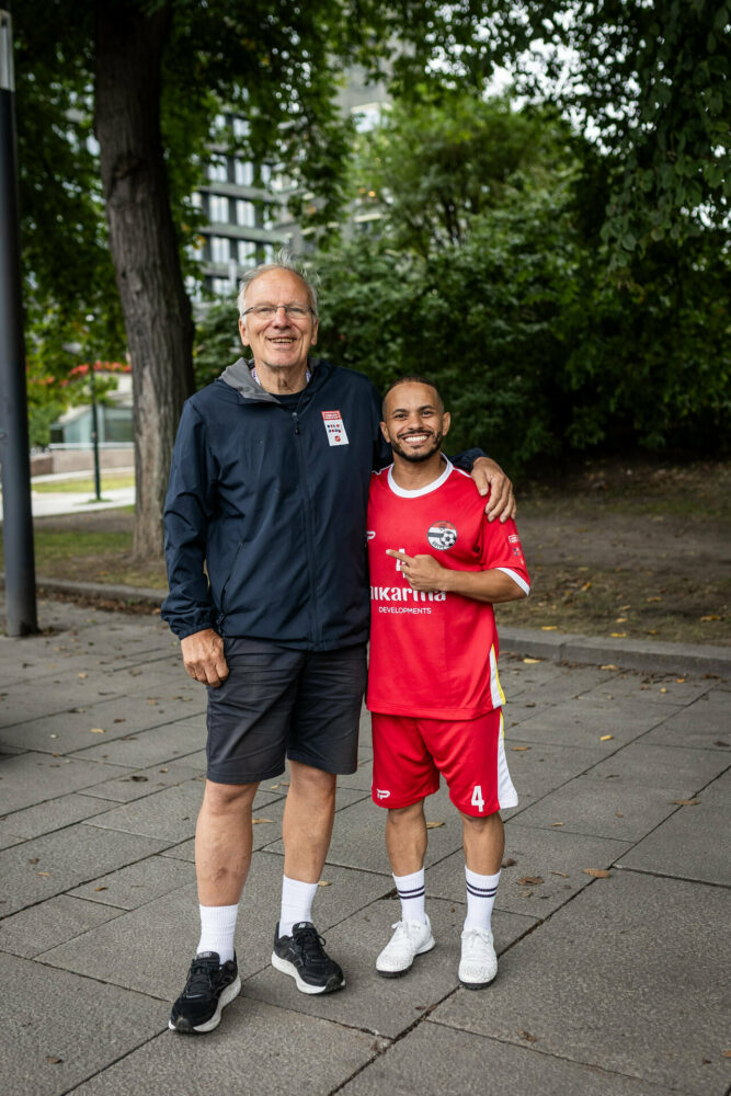 Foto: Terje Thodesen i blå HWC-jakke står med armen rundt en spiller som er mye lavere enn ham. Spilleren har på seg rød spillerdrakt.