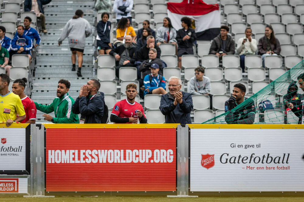 Foto: Terje Thodesen står rett utenfor banen og klapper under en kamp. Andre spillere som ikke er på banen da står ved siden av og roper og klapper. I bakgrunnen sees tribunen med andre publikummere.
