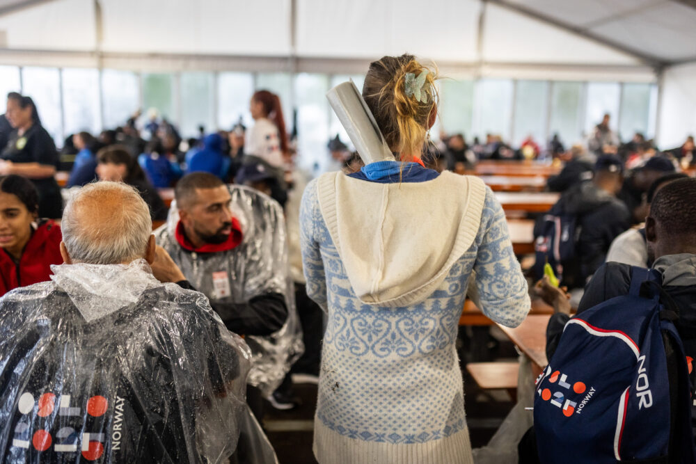 Foto: Silje Marie går innover i matteltet, bildet er tatt bakgfra. En rull med søppelsekker stikker opp av den hvite og blå strikkejakka. Rundt henne sitter spillere og spiser, noen av dem med gjennomsiktig regnponcho.