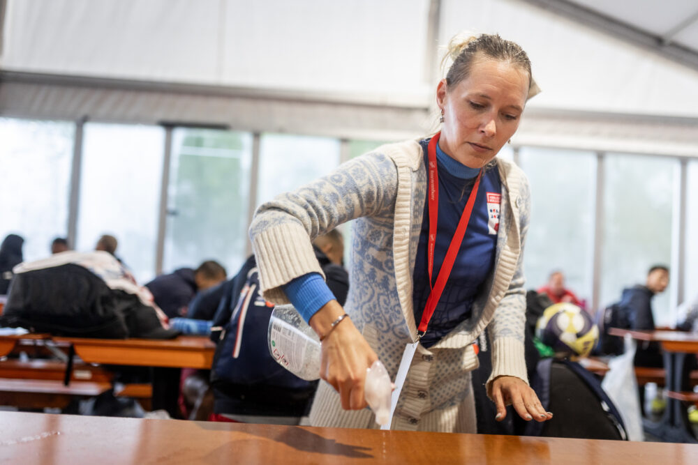 Foto: Silje Marie I hvit og lyseblå strikkejakke og lyst hår i hestehale sprayer rengjøringsspray på et trebord. I bakgrunnen sees spillere uskarpt.