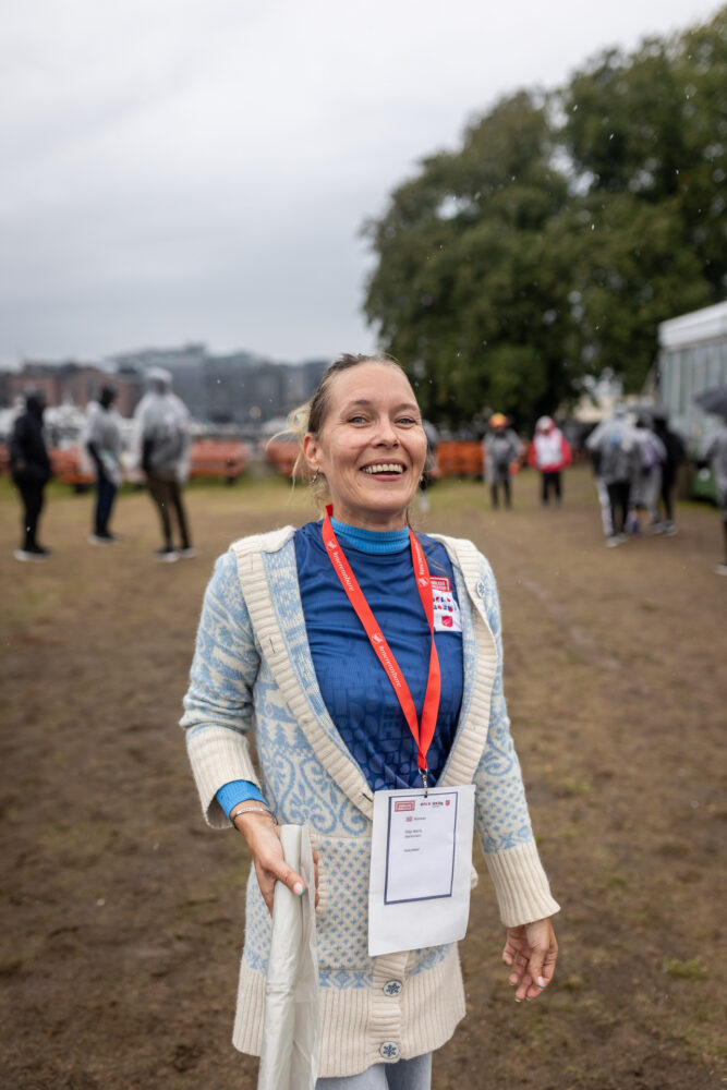 Foto: Silje Marie står og smiler mot kamera. Hun har på seg blå t-skjorte med HWC-logo og en hvit og blå strikkejakke over. I hånden holder hun en rull med søppelposer. I bakgrunnen skimtes fotballspillere i regnponcho.