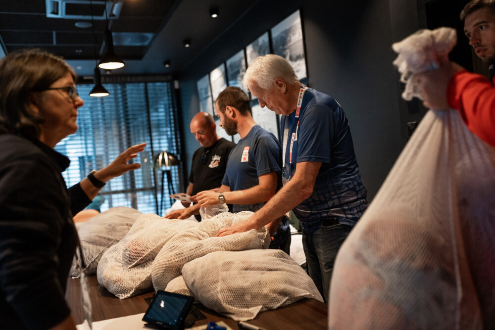Foto: tre menn, deriblant frivillig Steinar står foran et bord med hvite sekker med klær og sorterer. En kvinne i svart jakke snakker med en spiller som er utenfor bildet, bare en hånd som holder en sekk synes.